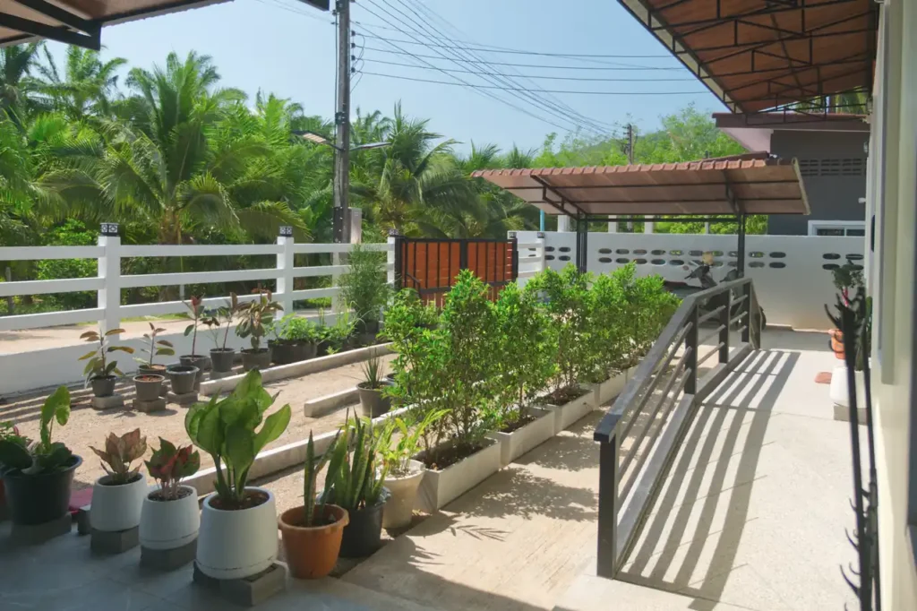Front garden and terrace of Baan Bahng Can Do Krabi homestay with planted raised beds, potted tropical plants, covered porch, and open gate