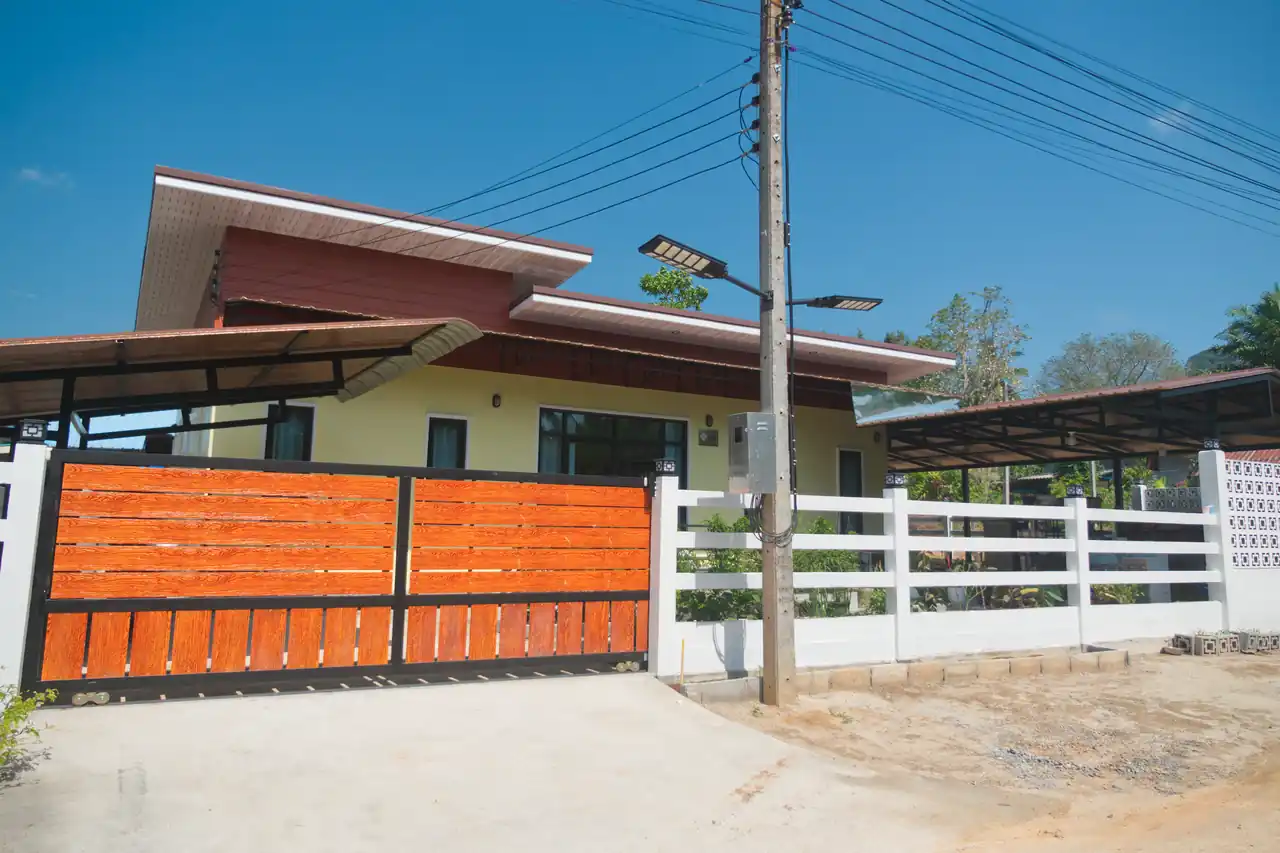 Gated entrance and exterior of Baan Bahng Can Do homestay in Nong Thale, Krabi, with timber-panel sliding gate, white perimeter fence, and covered carport