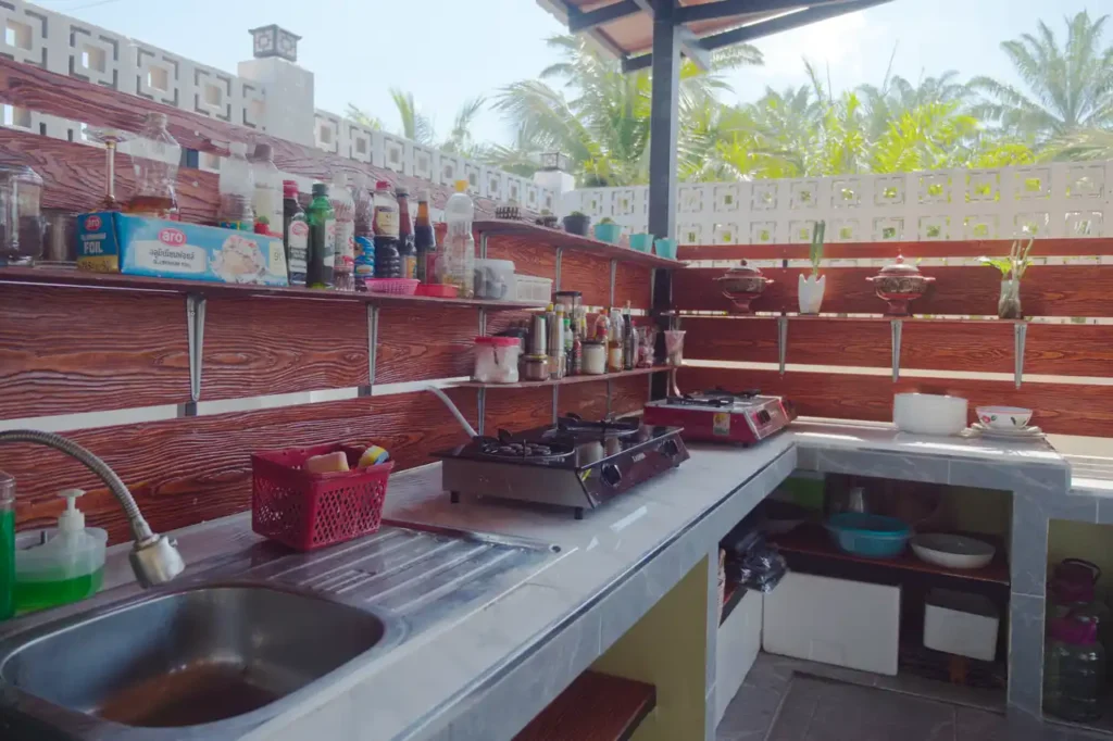 Outdoor Thai cooking kitchen at Baan Bahng Can Do Krabi homestay with gas hob, sink, fully stocked spice and sauce shelves, and palm tree views beyond the perimeter wall
