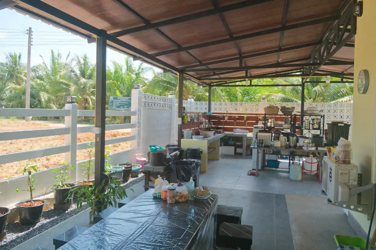 Wide view of the covered outdoor kitchen and cooking terrace at Baan Bahng Can Do Krabi homestay, with prep benches, cooking equipment, and open countryside beyond the white fence