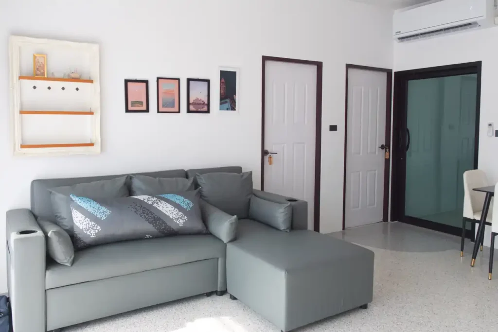 Shared living room at Baan Bahng Can Do Krabi homestay with L-shaped grey sofa, wall shelf, travel photo prints, air conditioning, and doors to guest bedrooms